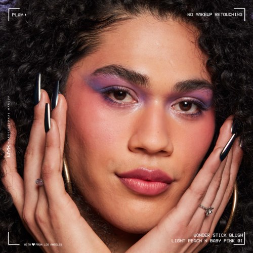 Vrouw met kleurrijke oogmake-up, krullend haar en ringen, raakt haar gezicht voorzichtig aan.