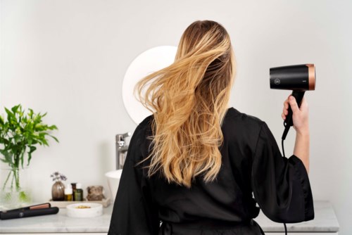 A woman with long blonde hair dries her hair at a vanity mirror, with a plant and beauty items nearby.