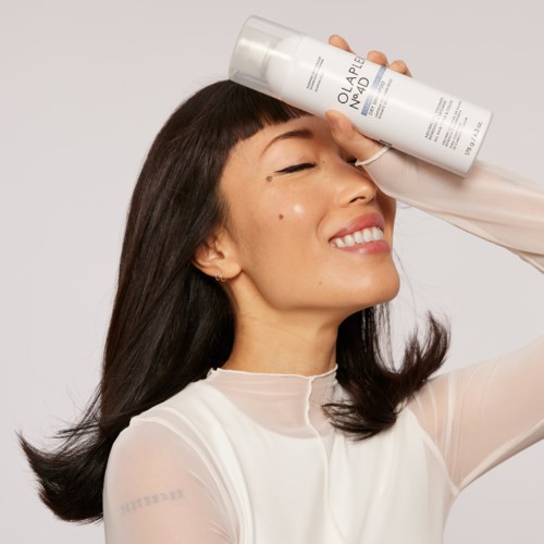 Een vrouw met donker haar glimlacht en houdt een witte fles huidverzorgingsproduct boven haar voorhoofd.