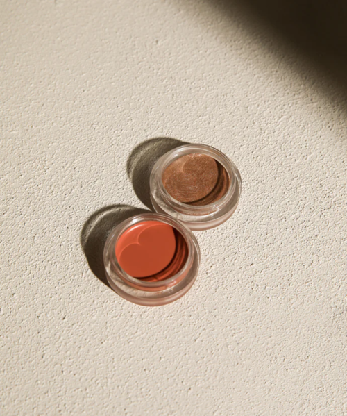 Twee kleine potsjes met perzik en bruin make-up of crème, geplaatst op een lichte, gestructureerde ondergrond.