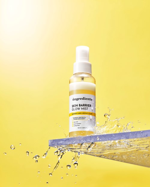Sumutinpullo nimeltä 'Skin Barrier Glow Mist' kelluu lautapinnalla, veden roiskeita ympärillä, keltainen tausta.