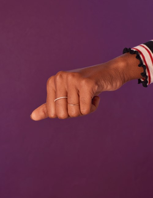 En hand med ringar pekar åt vänster mot en lila bakgrund, iklädd en randig ärm med svarta rufflekanter.