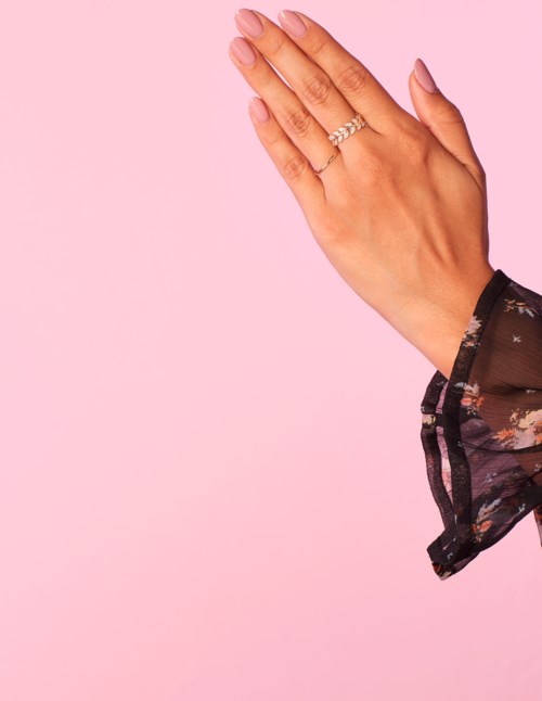 En kvinnas hand med ringar och rosa naglar, håller sin andra hand mot en rosa bakgrund, med del av ett blommigt ärm synligt.