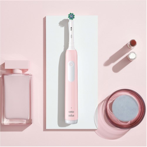 An electric toothbrush with a pink handle, two small bottles, a glass container, and a pink box, all on a pink background.