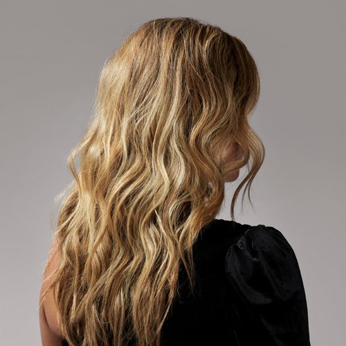 Eine Frau mit langen, gewellten blonden Haaren, trägt eine schwarze Oberbekleidung, seitlich betrachtet.