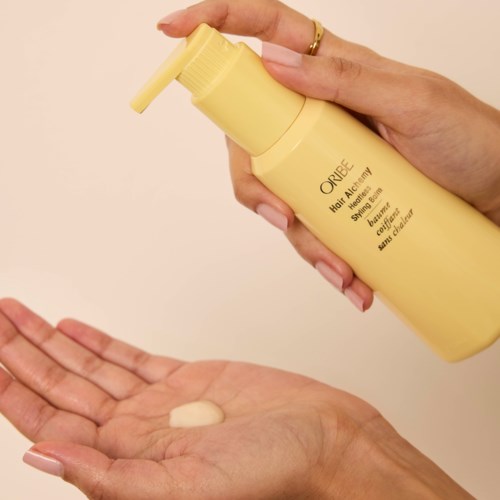 Iemand gebruikt een gele fles Oribe Heat Styling Spray, houdt het boven haar hand met een dot product.