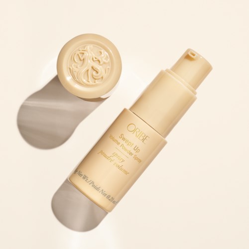 Een beige spuitfles met 'Oribe'-label en een decoratief deksel, met schaduw op een lichte achtergrond.