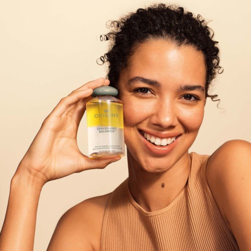 A woman with curly hair smiling, holding a bottle labeled 'Origins Checks and Balances' close to her face.