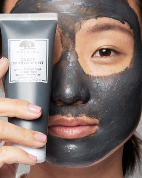 Eine Frau trägt eine schwarze Gesichtsmaske auf, während sie eine Tube von Origins hält. Nur ein Auge ist sichtbar.