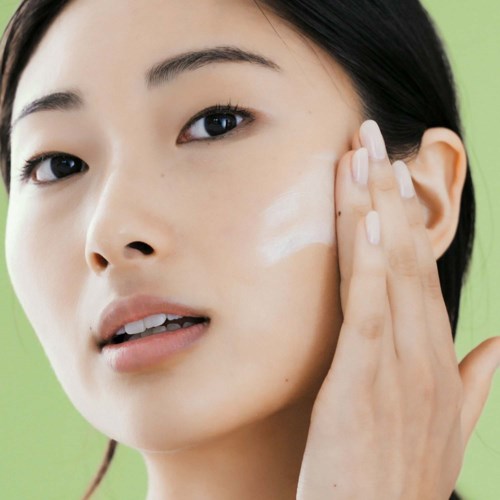 Eine Frau trägt Creme oder Lotion auf ihr Gesicht auf mit der Hand, mit grünem Hintergrund.