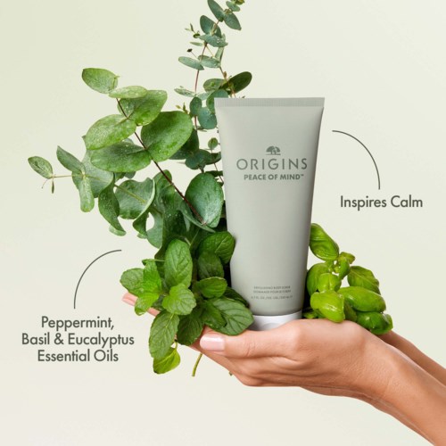 A hand holds a white tube labeled 'Origins Peace of Mind' with herbs, including peppermint, basil, eucalyptus, and green berries.