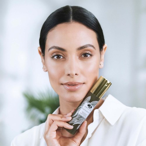 Eine Frau mit langen dunklen Haaren hält ein Hautpflege- oder Kosmetikprodukt mit schwarz-goldenem Design und lächelt sanft.