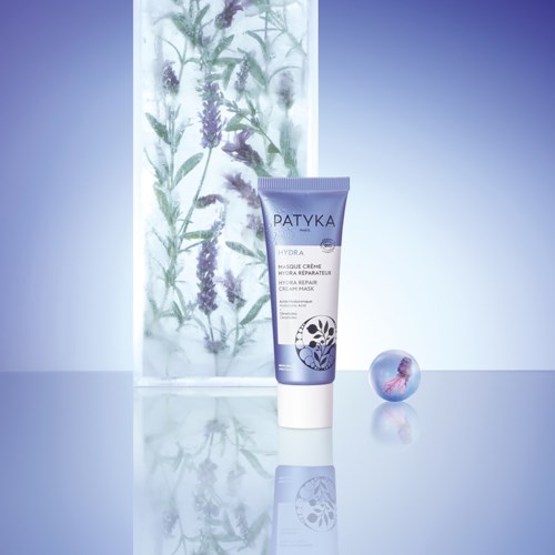 Eine lila Tube mit Patyka Hydra Reparerende Creme vor botanischem Hintergrund und einer Kristallkugel daneben.