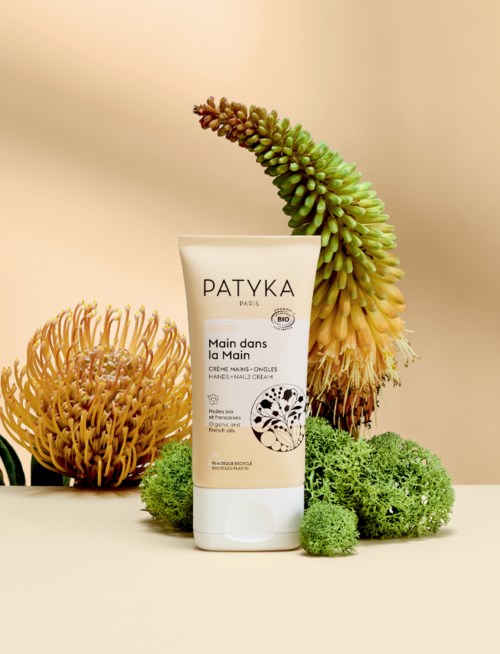 Eine Handcremetube mit natürlichen Pflanzen und Blumen im Hintergrund, beschriftet 'Patyka Main dans la Main.'