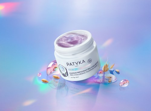 Ein Behälter mit 'PATYKA'-Aufschrift und violetter Creme, umgeben von Edelsteinen und getrockneten Blumen auf einer glänzenden Oberfläche.