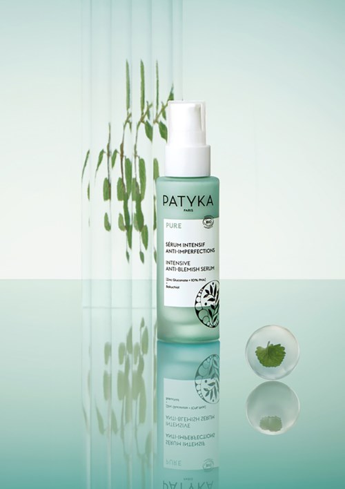 Eine Flasche Patyka Pure Anti-Imperfection Serum mit klarem, minimalistischen Design und weißer Pumpe auf reflektierender Oberfläche.