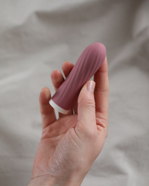 Eine Person hält einen lilafarbenen, penisförmigen Gegenstand, die Finger greifen ihn. Der Hintergrund ist hell und schlicht.