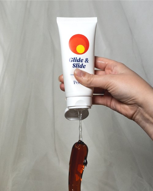 Eine Hand hält eine weiße Tube mit einem bunten Logo, aus der braune Gel herausquillt. Der Hintergrund ist schlicht.