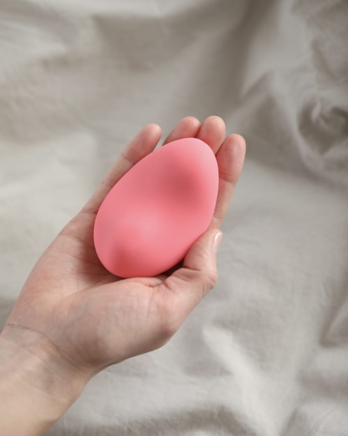 Eine Hand hält einen großen, rosa, eiförmigen Make-up-Schwamm vor einem grauen Stoffhintergrund.