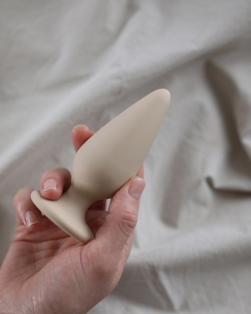 Eine Hand hält ein beiges, glattes, eiförmiges Objekt mit flacher Basis gegen einen geriffelten hellfarbenen Stoff.