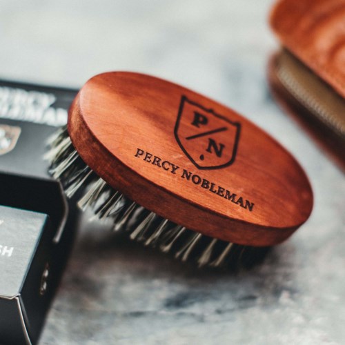 A wooden grooming brush with the Percy Nobleman logo and metal bristles on a surface.