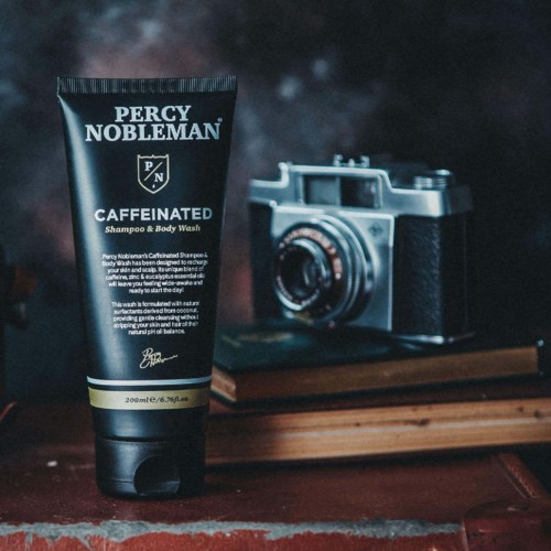 A black tube of Percy Nobleman Caffeinated shampoo and body wash is placed on a wooden surface with a camera nearby.
