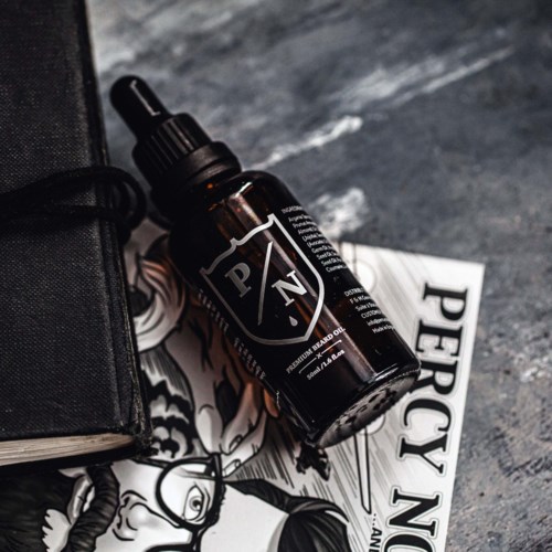 A dark bottle of beard oil with a spray cap, placed on a printed paper, with part of a black bag nearby.
