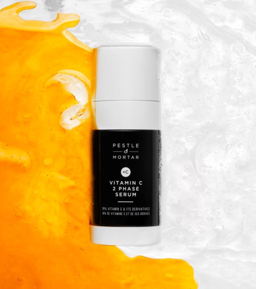 Een zwart-wit flesje met 'Vitamin C 2 Phase Serum' ligt op een oppervlak met oranje en witte vloeistoffen.