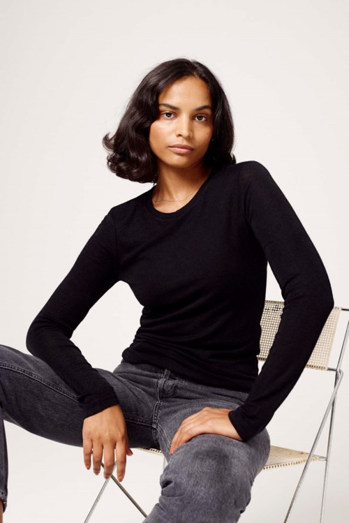 En kvinna i svart tröja och grå jeans sitter på en vit stol mot en ljus bakgrund.
