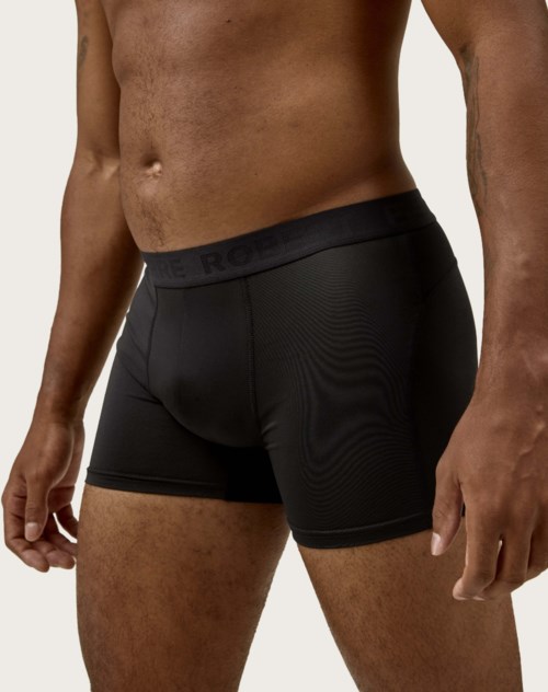 Ein Mann trägt schwarze Boxershorts mit sichtbarem Bund. Sein Oberkörper und die Arme sind ebenfalls sichtbar.