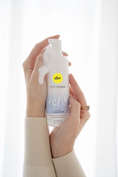 En person håller en vit flaska Pjur Toy Foam rengöringsskum, med skum som rinner ut från toppen.