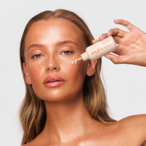 Een vrouw met lang haar brengt lotion aan op haar gezicht. Ze heeft een neutrale uitdrukking en kijkt naar de camera. De achtergrond is wit.