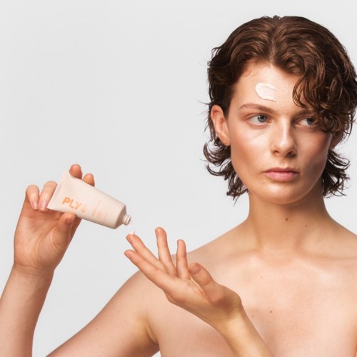 Een vrouw brengt lotion aan op haar hand, met een pleister op haar voorhoofd, tegen een eenvoudige achtergrond.