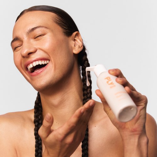 Een lachend persoon met lange vlechten brengt lotion aan op het gezicht vanuit een witte fles.