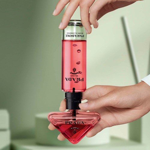 En person holder en Prada-parfymeflaske med rosa væske inni, opp ned, med en grønn uskarp bakgrunn.