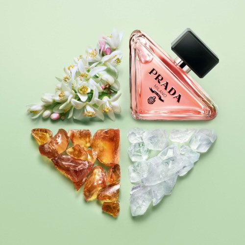 A floral bouquet, a triangular bottle of Prada perfume, amber stones, and ice cubes on a green background.