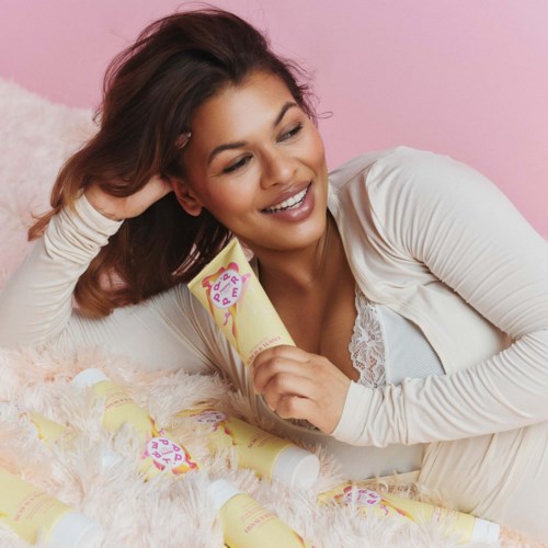 A smiling woman relaxing on a bed with multiple yellow bottles around her and holding one bottle.