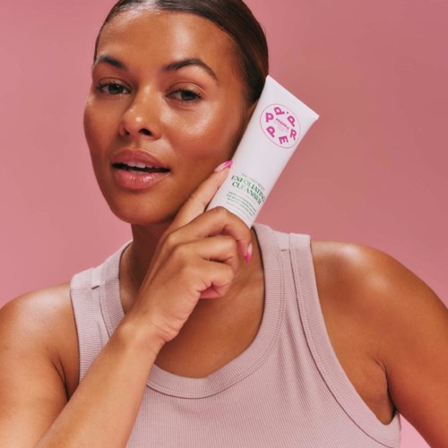 A woman with smooth skin holding a skincare product, with a pink background and a neutral expression.