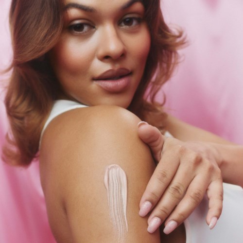 A woman applying white cream on her shoulder, with a pink background and soft lighting.