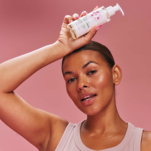 A woman with a pink background holds a small bottle above her head.
