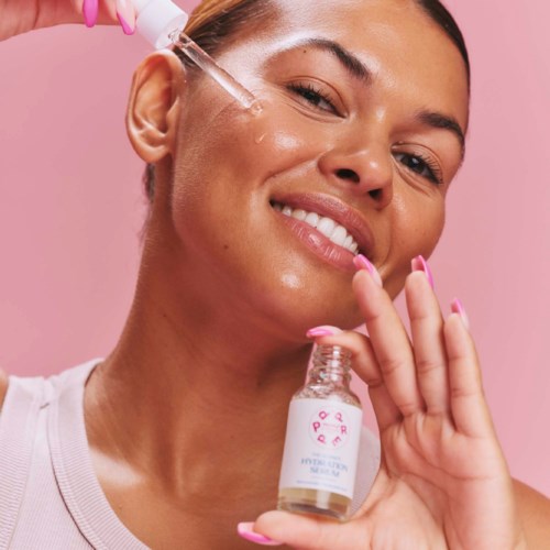 A woman applies skincare serum with a dropper, smiling in front of a pink background, holding a small bottle.