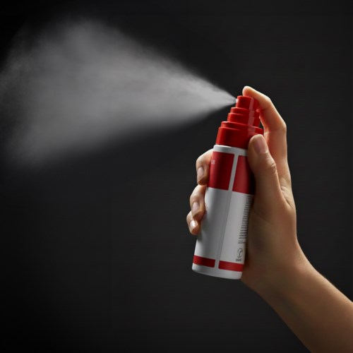 En hånd sprayer ut en hvit tåke fra en rød og hvit aerosolkanna mot en svart bakgrunn.