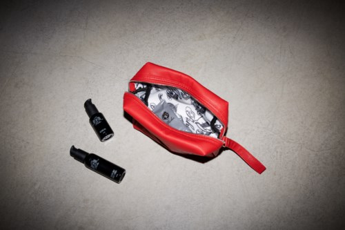 A red makeup bag with black and white items inside, and two small black bottles outside it on a grey surface.