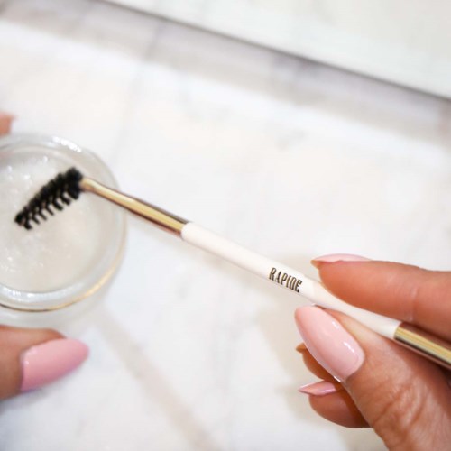 A hand holds a white and gold eyelash brush stick, cleaning a glass with water. The background is light and blurry.