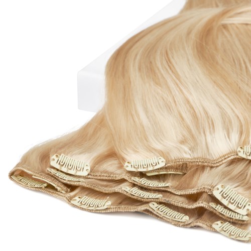 En blonder peruk med hårklämmor ligger på en vit yta bredvid en vit låda.