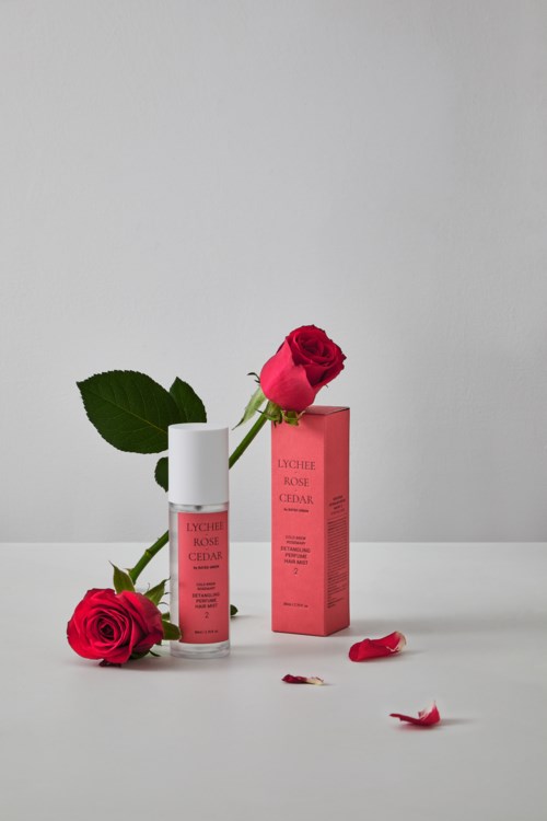 A white bottle with pink label, pink box, and two red roses on a white surface with petals.