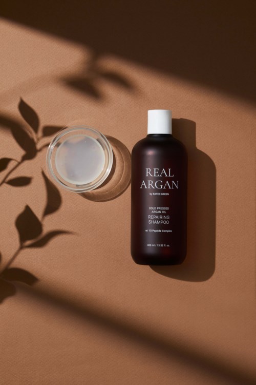 Een bruine fles met 'Real Argan' en een open container met witte crème, schaduwen op een beige achtergrond.