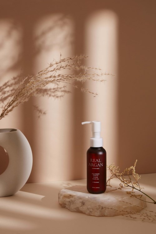 A bottle of real argan oil on a stone, with a vase and dried plants, in warm light and shadows.