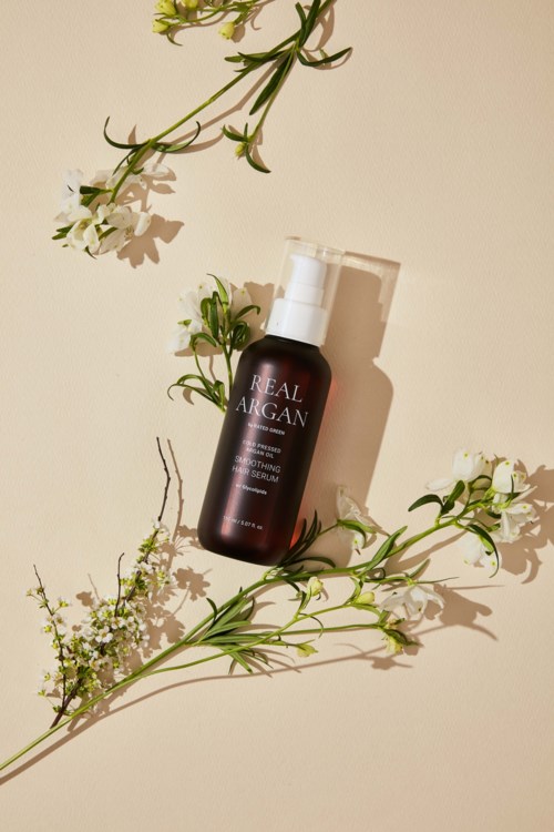 Een donkere fles met 'Real Argan' erop, omgeven door kersenbloesem en groene bladeren op een lichte achtergrond.