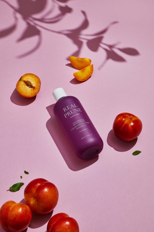 Een paarse fles met 'Real Prune' op, met perziken, bladeren en oranje-zijde zaden op een roze achtergrond.